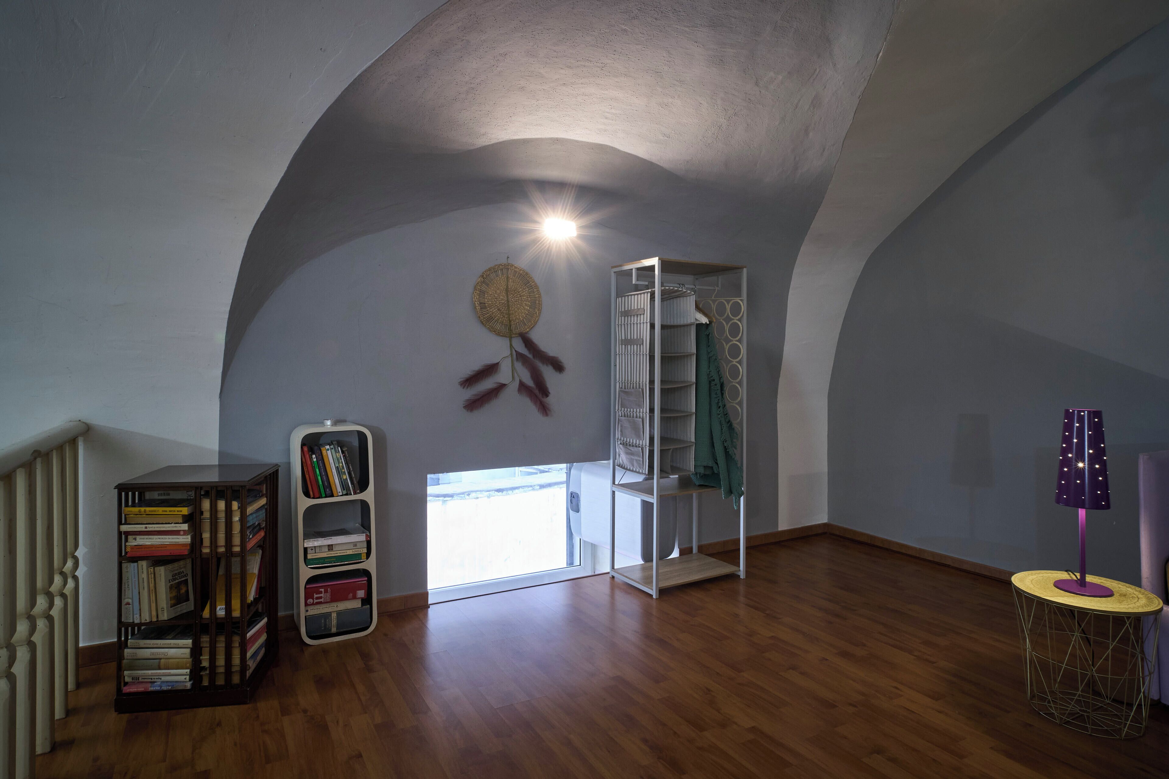 A room with wooden flooring, a bookshelf, a white shelving unit, and a lamp on a round table.