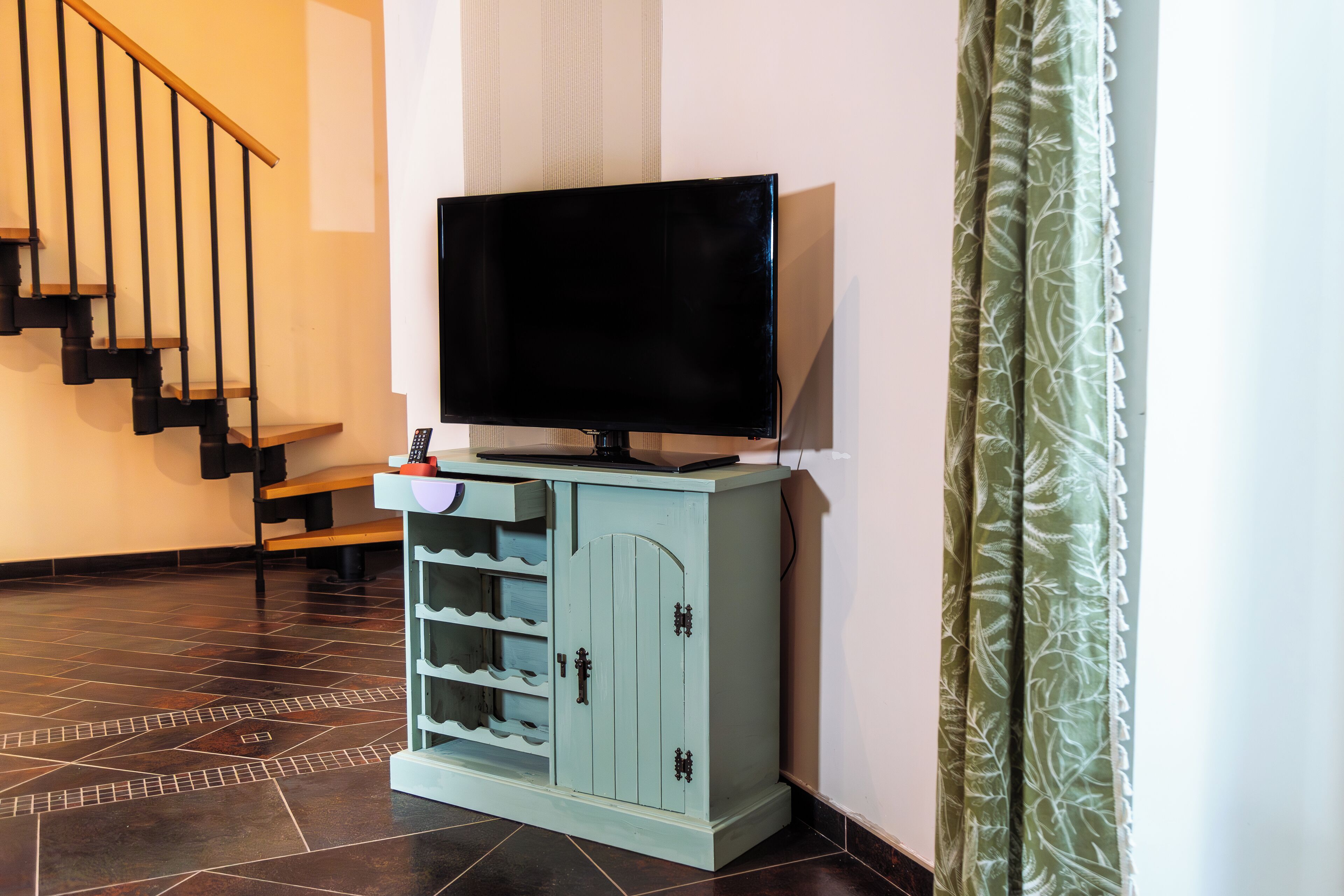 A living room with a television on a cabinet, a staircase, and a patterned curtain.
