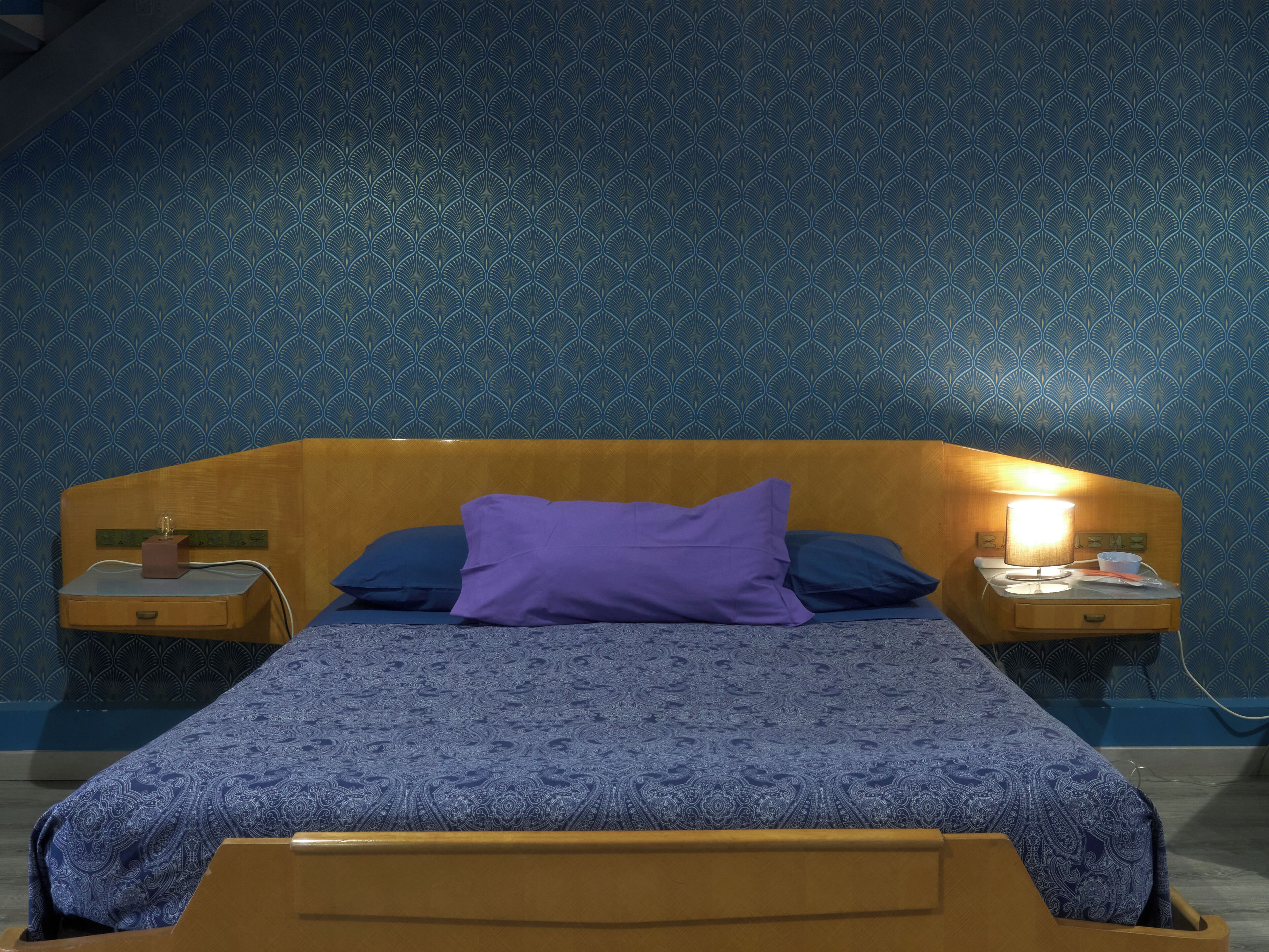A bedroom with a patterned blue wall, a wooden headboard, a bed with a blue patterned bedspread, two nightstands with lamps, and a purple pillow.