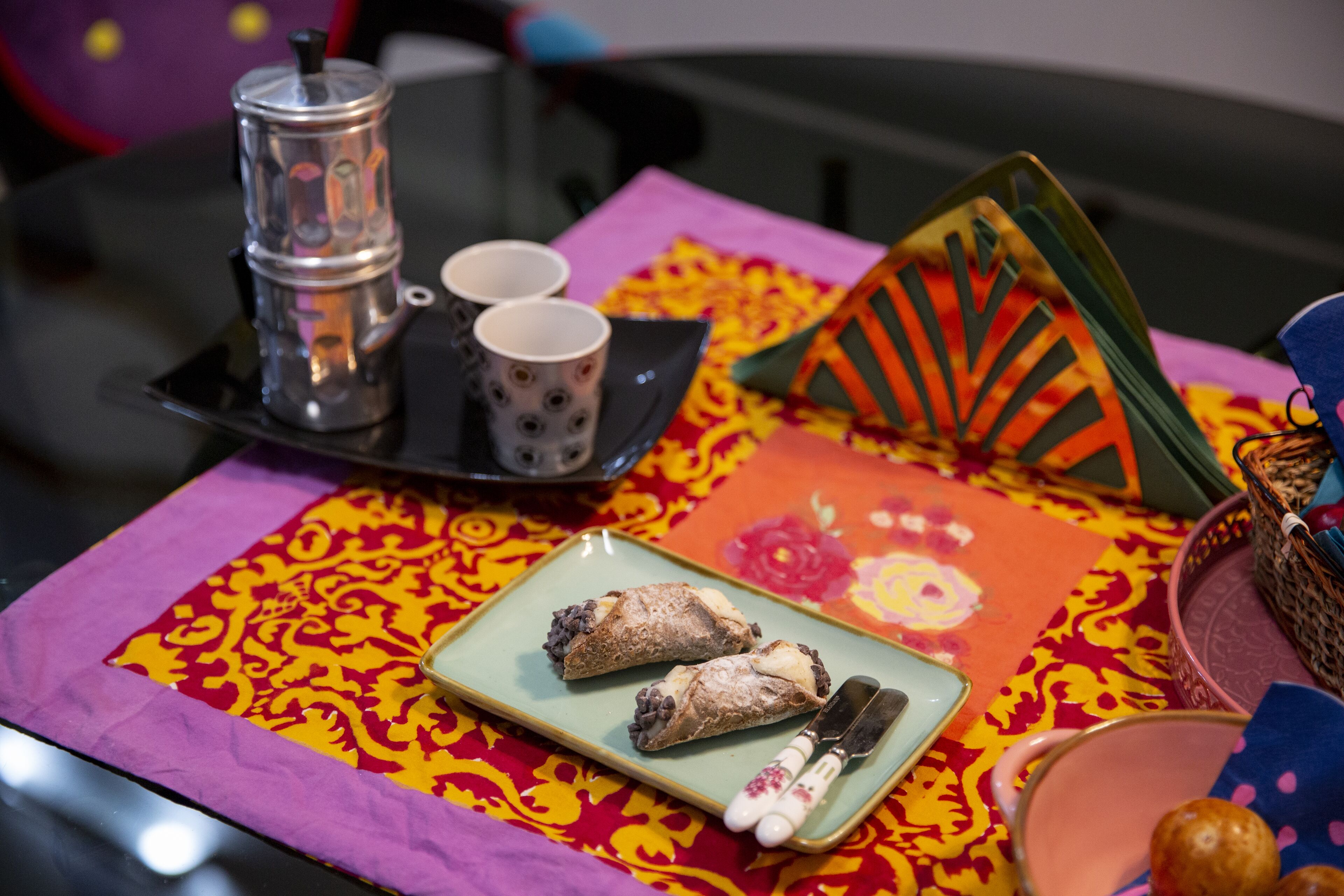 A table with a colorful tablecloth, a plate with pastries, a teapot, and cups.