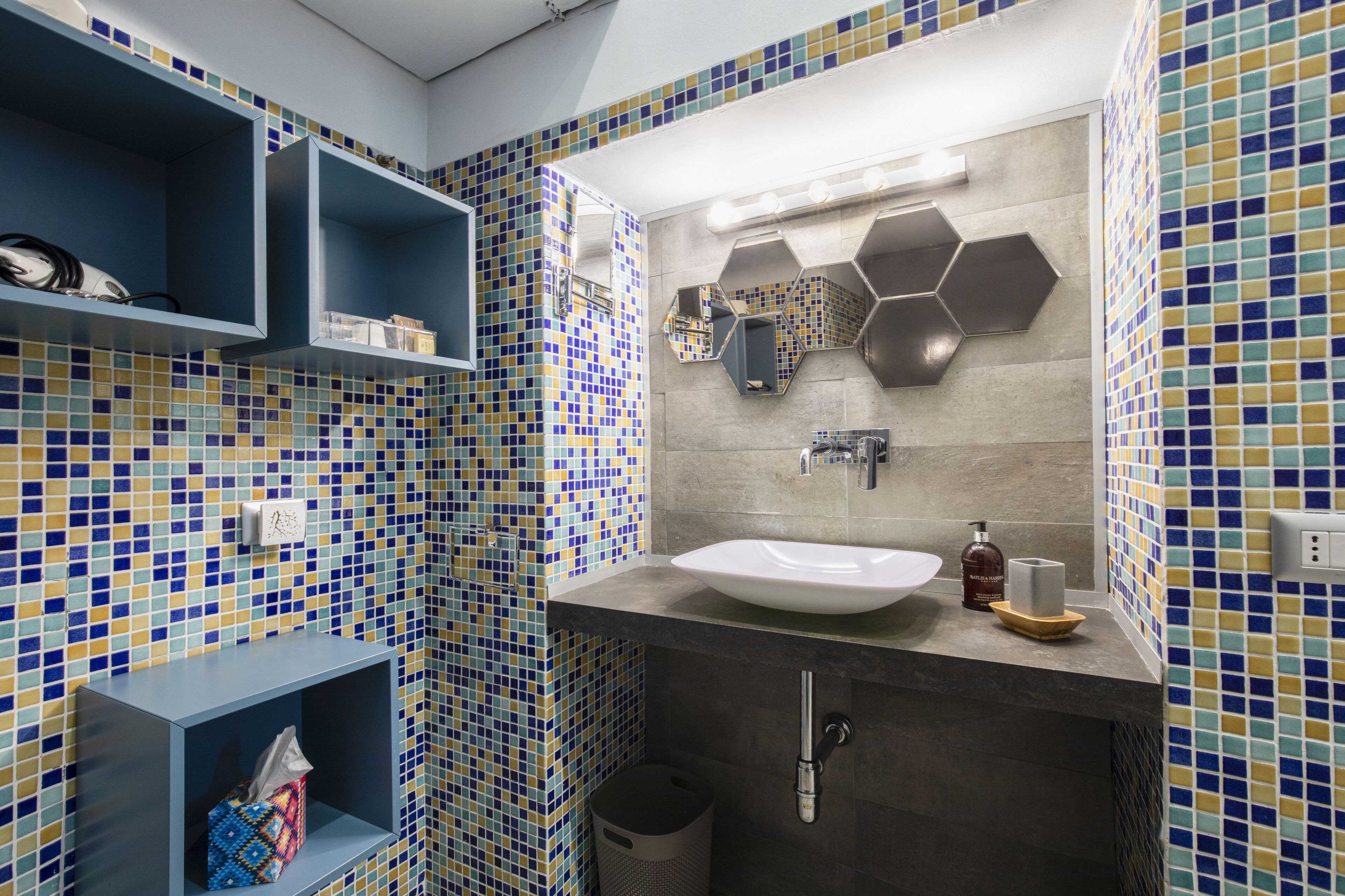 A bathroom with a tiled mosaic wall, a white sink, and a mirror. There are shelves with toiletries and a trash bin.