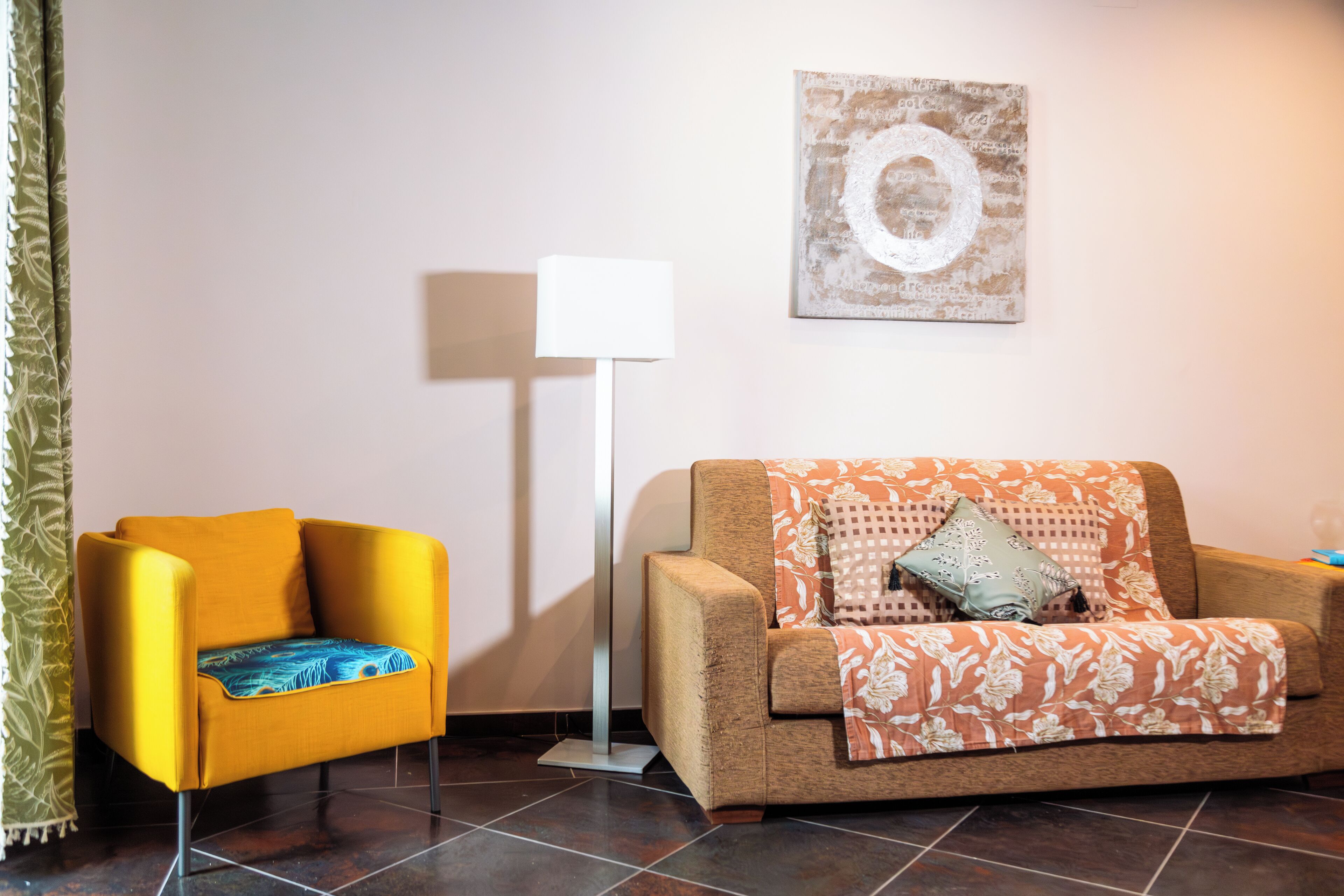 A room with a yellow armchair, a brown sofa with a floral throw, a floor lamp, and a wall with a circular design.