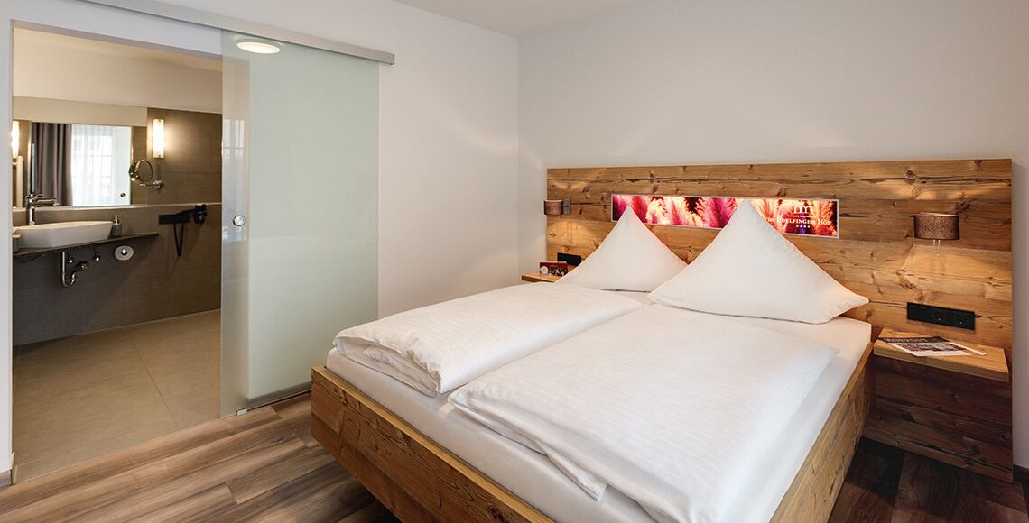 A modern hotel room with a wooden headboard, a large bed with white linens, a wooden nightstand, and a bathroom visible through an open door.
