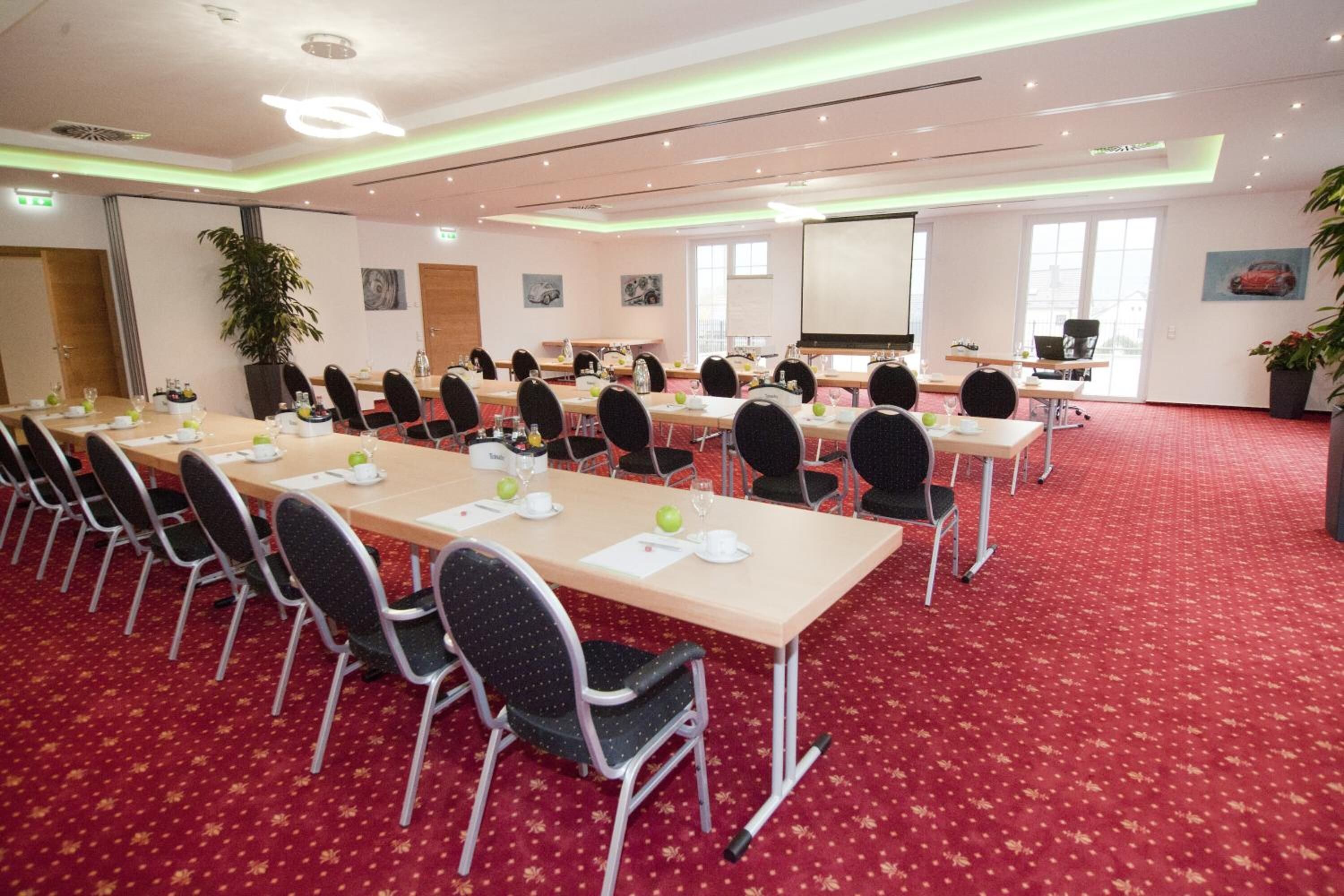 Une salle de conférence avec de longues tables et des chaises, un écran de projection et un sol recouvert d’un tapis rouge.