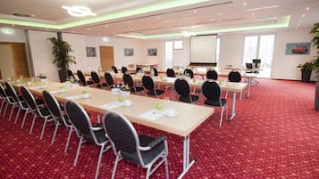 Une salle de conférence avec de longues tables et des chaises, un écran de projection et un sol recouvert d’un tapis rouge.