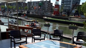 A glass-top table with wooden legs on a deck overlooking a canal with boats and buildings in the background. - Rooms on Water (Rotterdam)