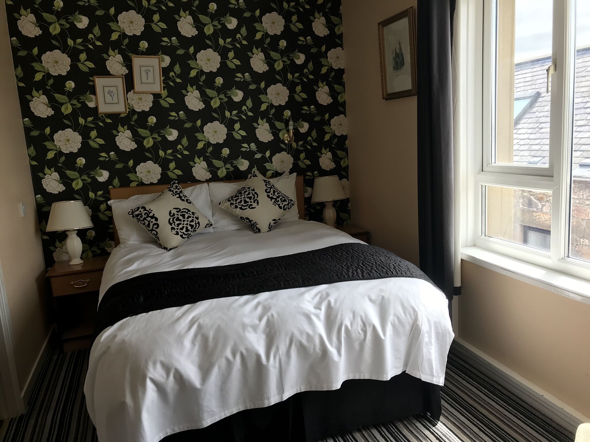 A neatly made bed with white and black bedding, a floral wallpaper, and a window with curtains.