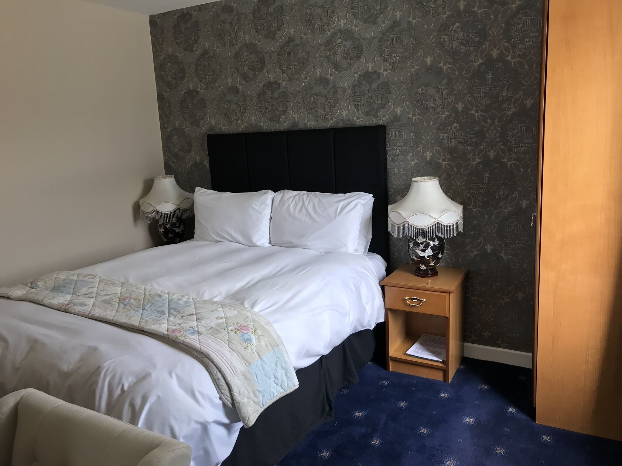 A neatly made bed with white linens, a dark headboard, and a blue patterned carpet.