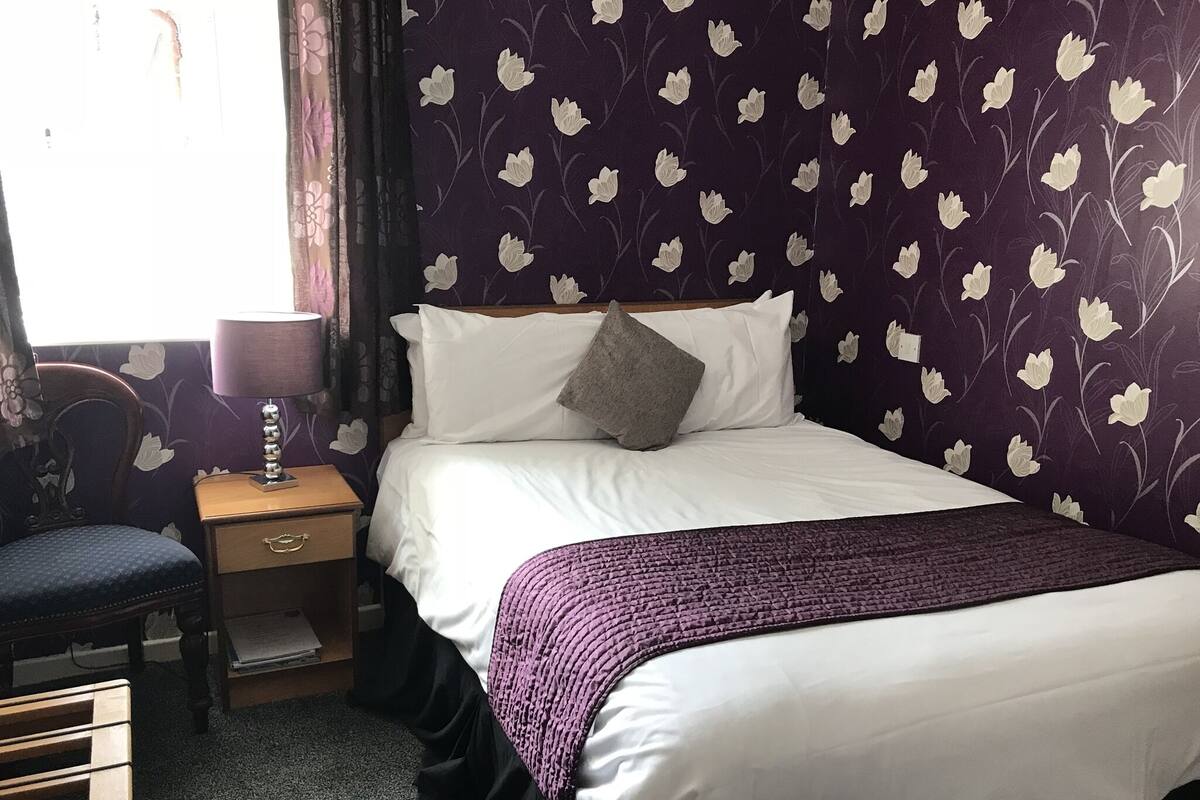 A neatly made bed with white linens and a purple blanket, a wooden nightstand with a lamp, a chair, and a wall with a floral pattern.