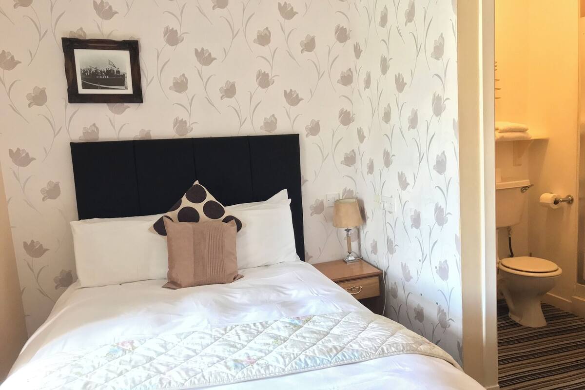 A neatly made bed with a quilt, a nightstand with a lamp, a framed picture on the wall, and a toilet visible through an open door.