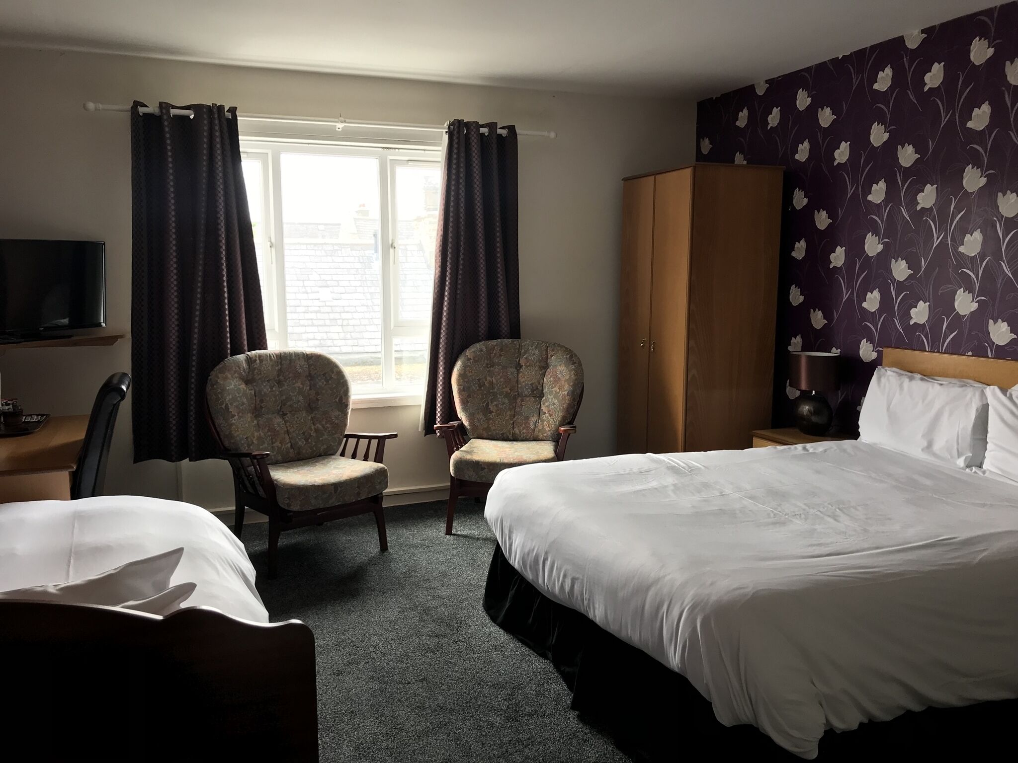 A hotel room with two single beds, a large wardrobe, a television, and two armchairs.