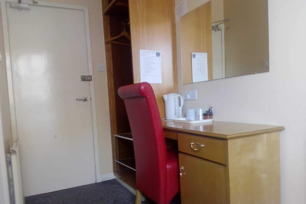 A hotel room with a wooden desk, a red chair, a white bed, a mirror, and a door.