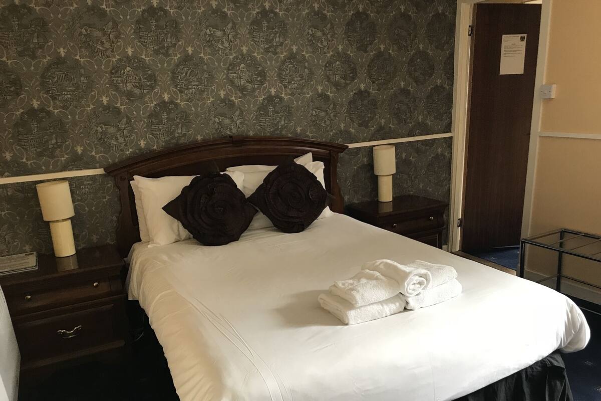 A neatly made bed with white linens and dark decorative pillows, a folded towel on the bed, and a dark wooden headboard.