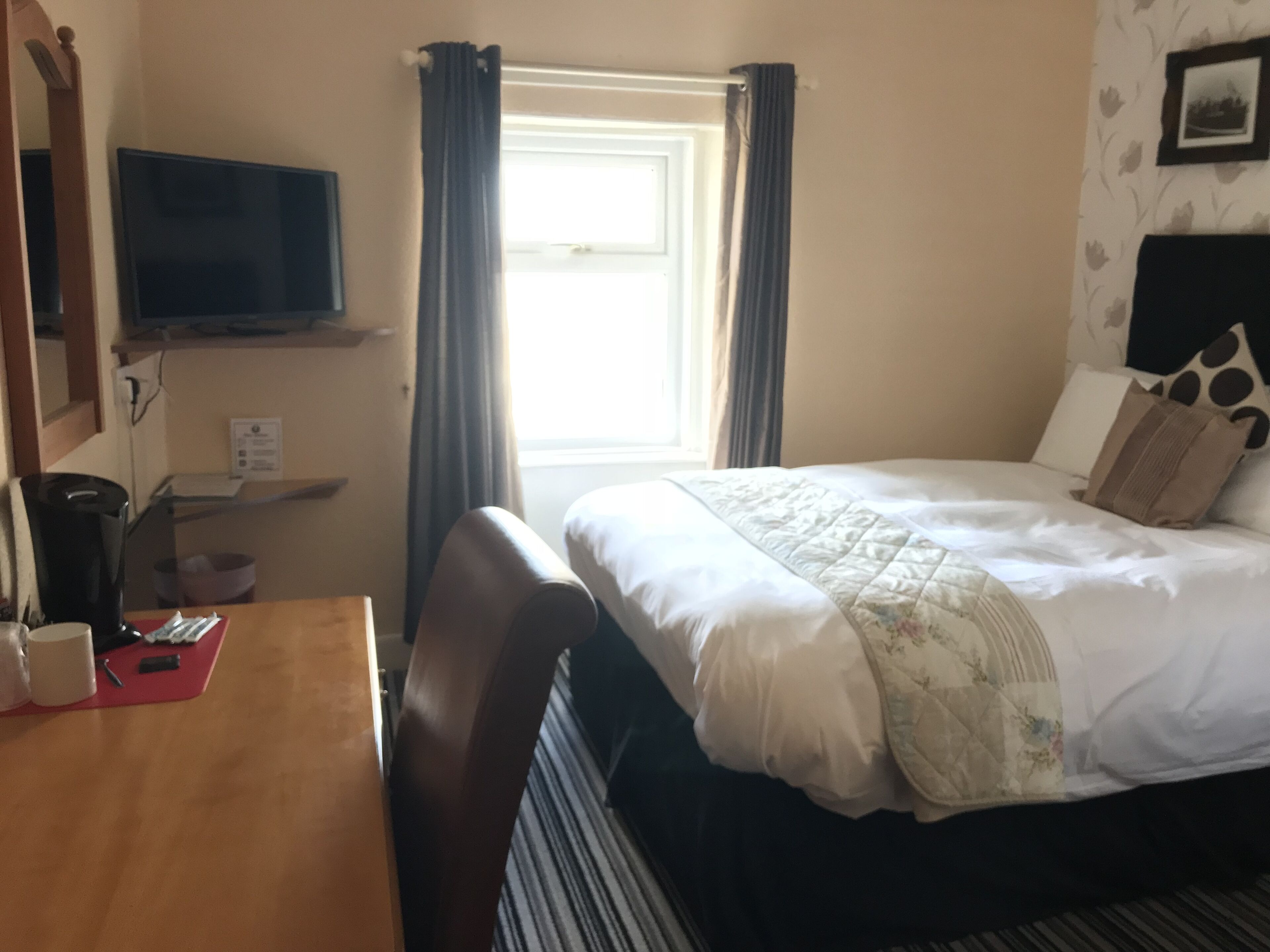 A hotel room with a bed, a TV, a desk with a coffee maker, and a window with curtains.