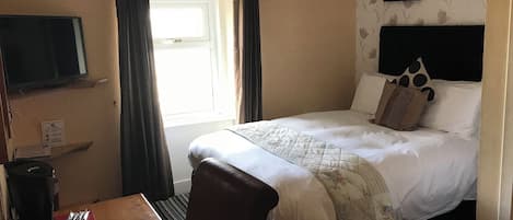 A hotel room with a bed, a TV, a table with a mug and a phone, and a window with curtains.