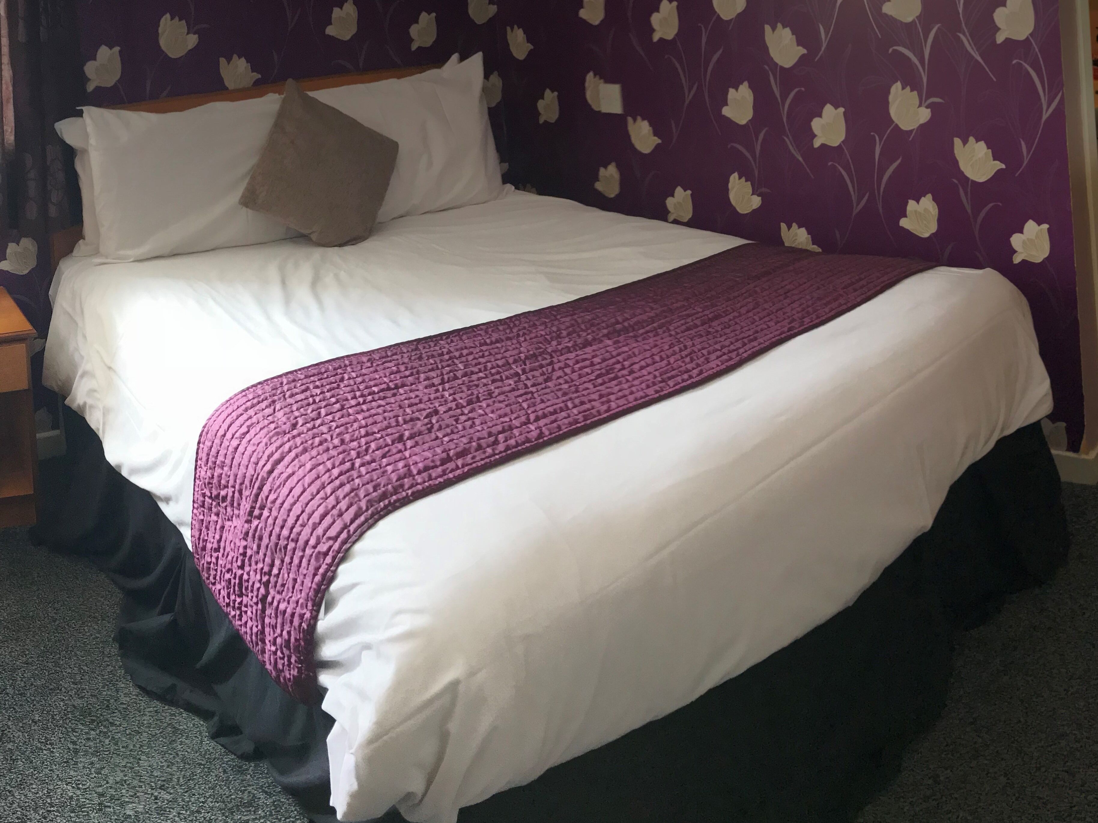 A neatly made bed with white linens and a purple blanket, set against a purple floral wallpaper.