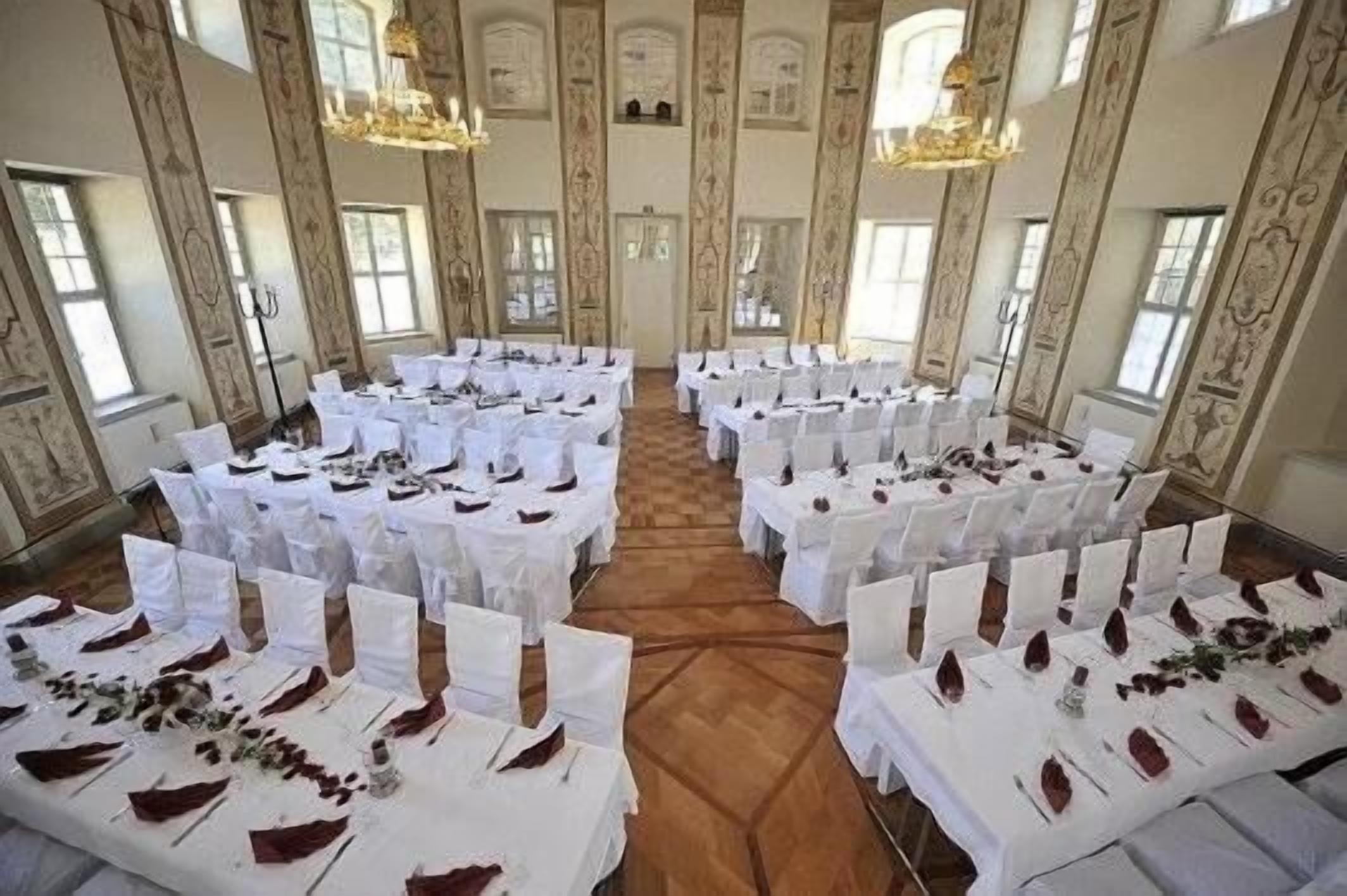 Uma sala de banquetes com várias mesas redondas montadas para um evento formal, com toalhas brancas e guardanapos vermelhos.
