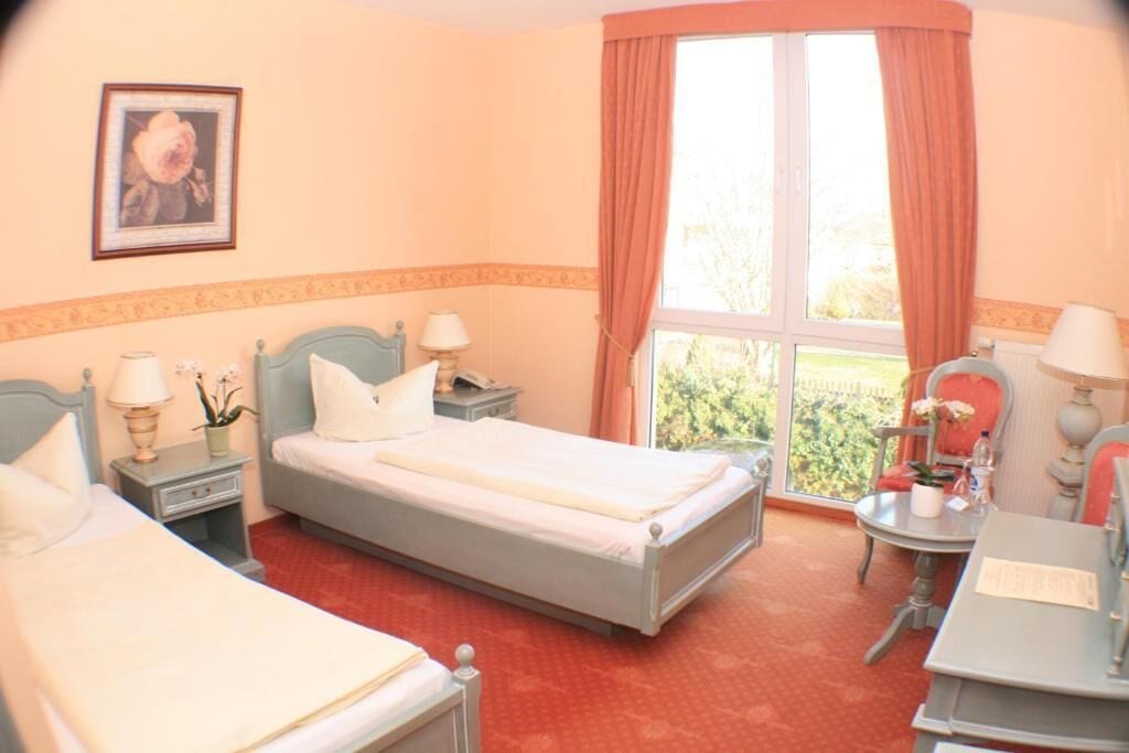 Quarto de hotel com duas camas, uma janela com cortinas, uma escrivaninha com cadeira e uma mesa pequena com abajur.