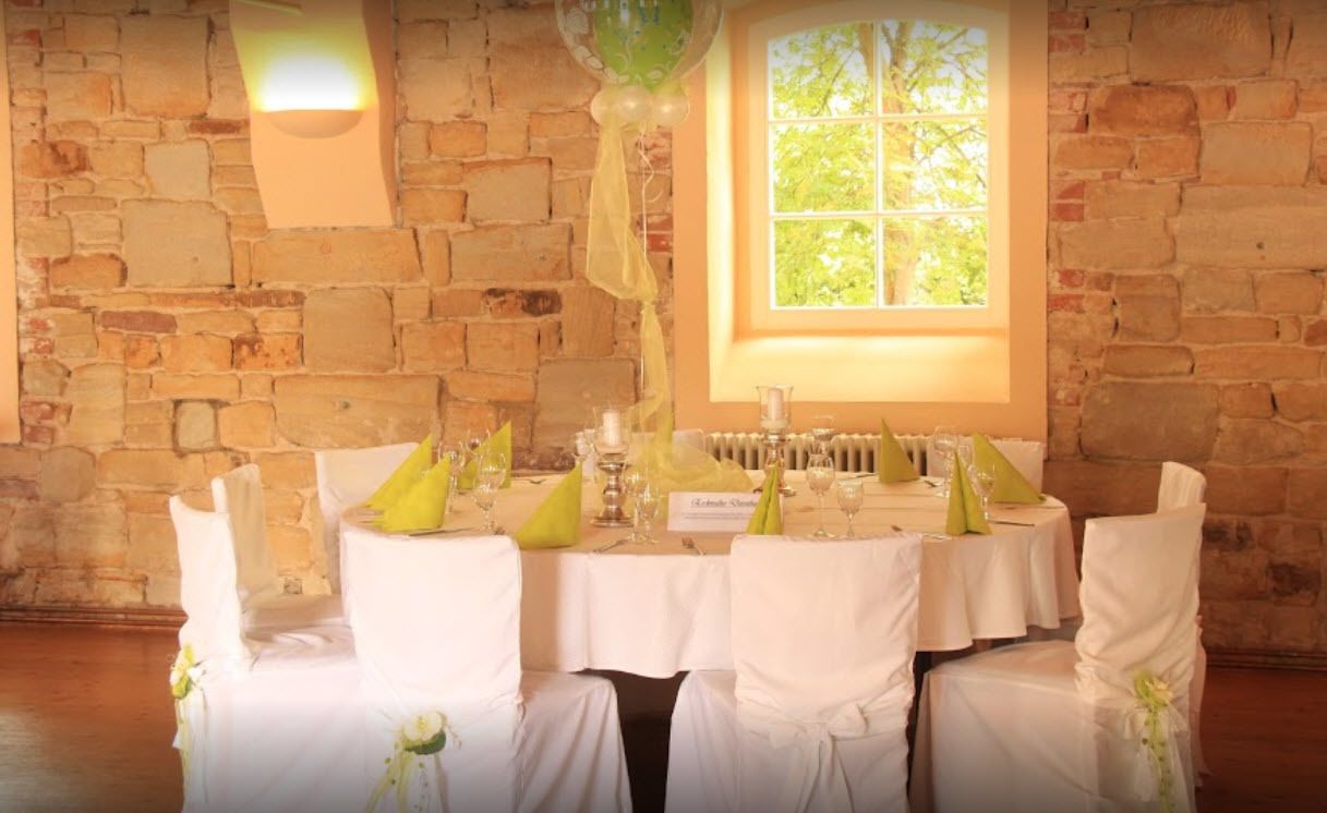 A round table set with white tablecloths, green napkins, and glassware, with a brick wall and a window in the background.