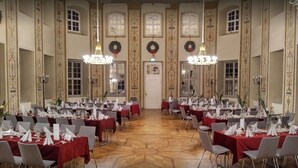 A banquet hall with multiple round tables set for dining, large chandeliers, and a high ceiling with decorative moldings.