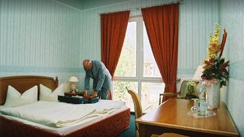 A hotel room with a bed, a wooden nightstand with a lamp, a dining table with chairs, and a window with curtains.
