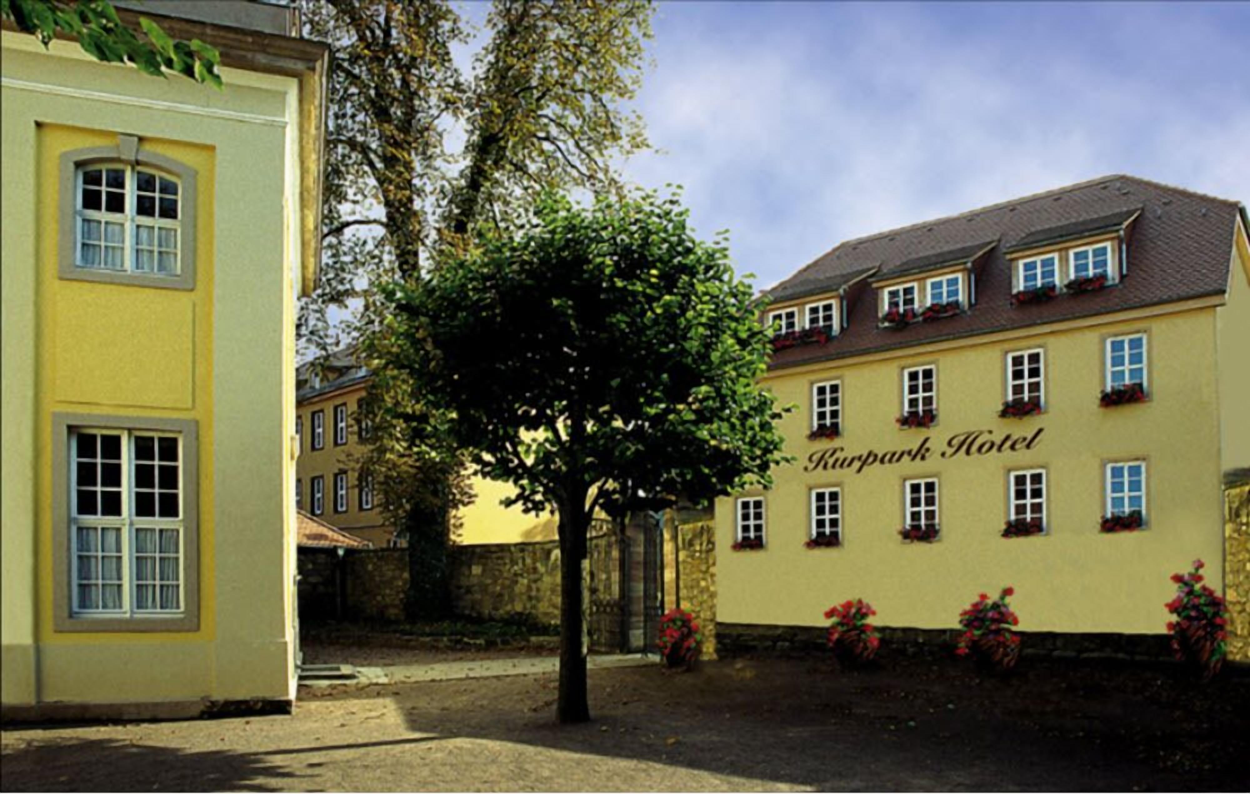 Un bâtiment jaune, portant l’inscription « Rurpark Hotel » sur sa façade, entouré d’arbres et d’un mur de pierre.