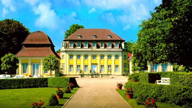 A large, symmetrical building with a central entrance, surrounded by well-maintained gardens and a clear sky.