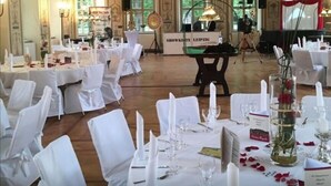 A banquet hall with high ceilings, ornate chandeliers, and large windows. Tables are set with white tablecloths and candles.