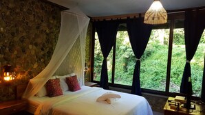 A bedroom with a stone wall, a bed with white bedding, a canopy, and a view of greenery through large windows. - Thansila Resort (Ranong)