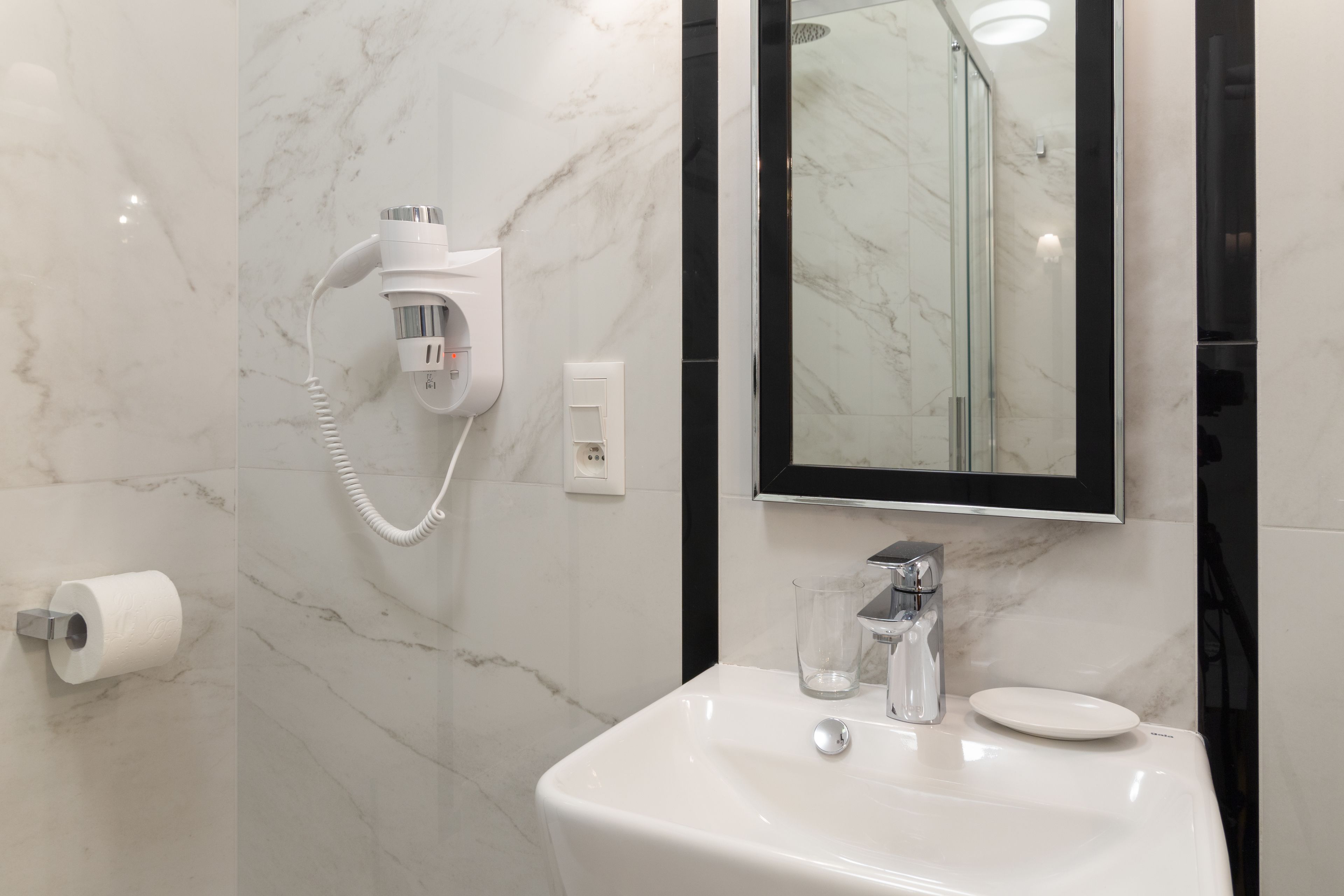 Un baño con pared de mármol, lavamanos blanco, cabina de ducha de vidrio y secador de pelo incorporado.