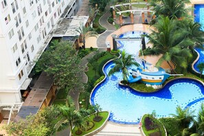 A swimming pool with a winding design, surrounded by palm trees and greenery, adjacent to a multi-story building. - Monochrome Style 2 Bedrooms at Kalibata City Apartment By Travelio (Jakarta)