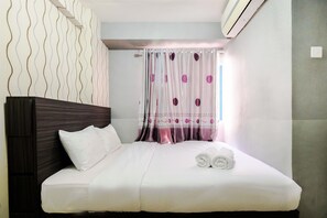 A neatly made bed with white linens and rolled towels, a dark wood headboard, and a window with patterned curtains.