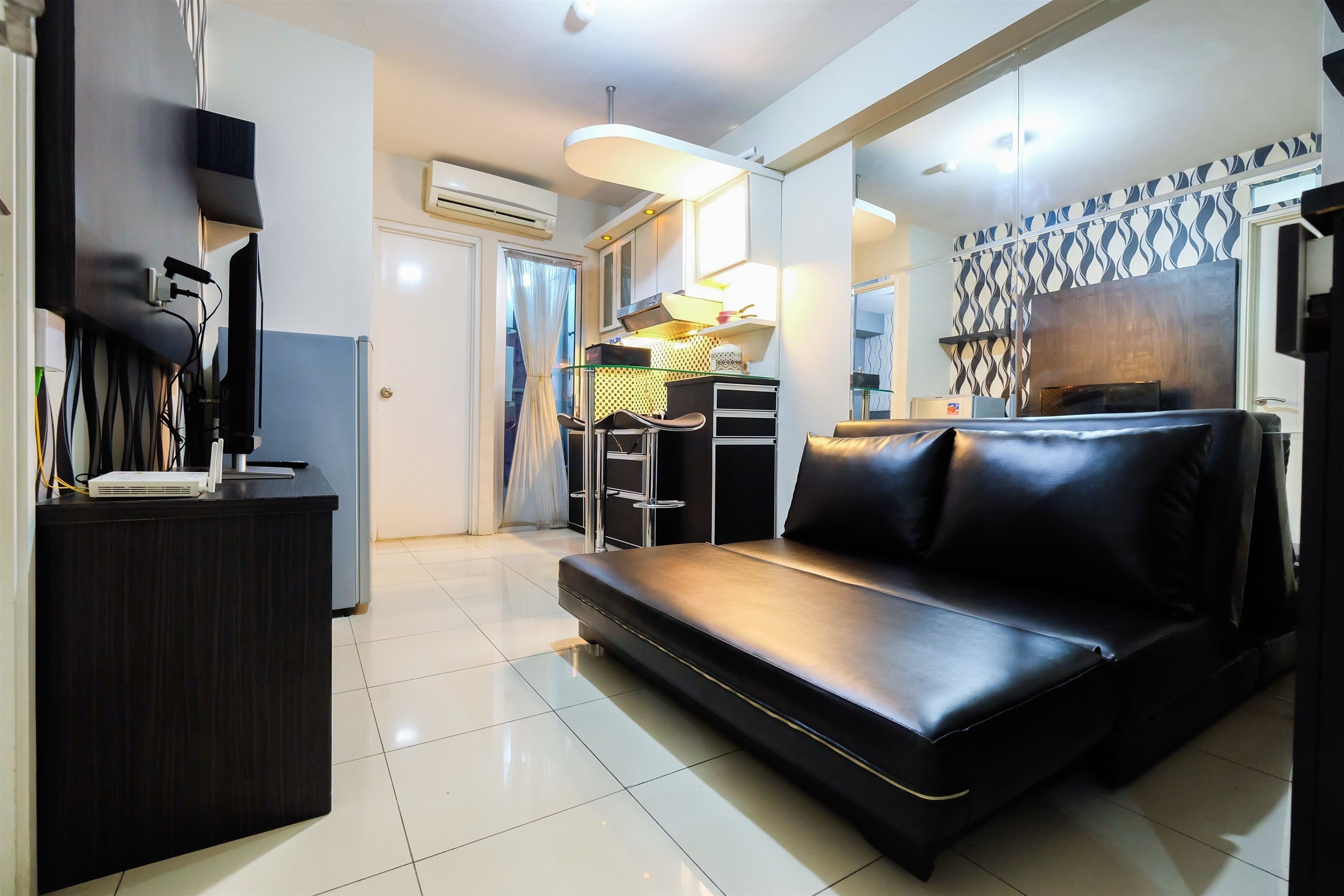Una sala moderna con un sofá de cuero negro, un escritorio de madera oscura y una pequeña área de cocina con microondas y una lámpara.