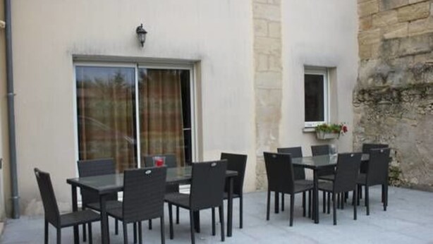 Une terrasse extérieure avec une table et des chaises en verre, un mur en pierre et une fenêtre ornée d’une plante.