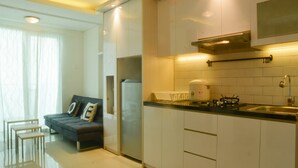 A modern kitchen with white cabinets, a black countertop, and a built-in stove. There is a dining area with a dark sofa and a small table. - 2 Bedrooms Woodland Park Apartment by Travelio (Jakarta)