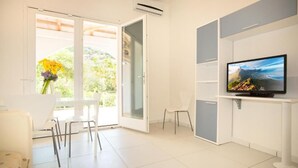 Una habitación moderna con una televisión sobre un mueble, un comedor con sillas y un ventanal corredizo que da a un balcón con vistas a zonas verdes.