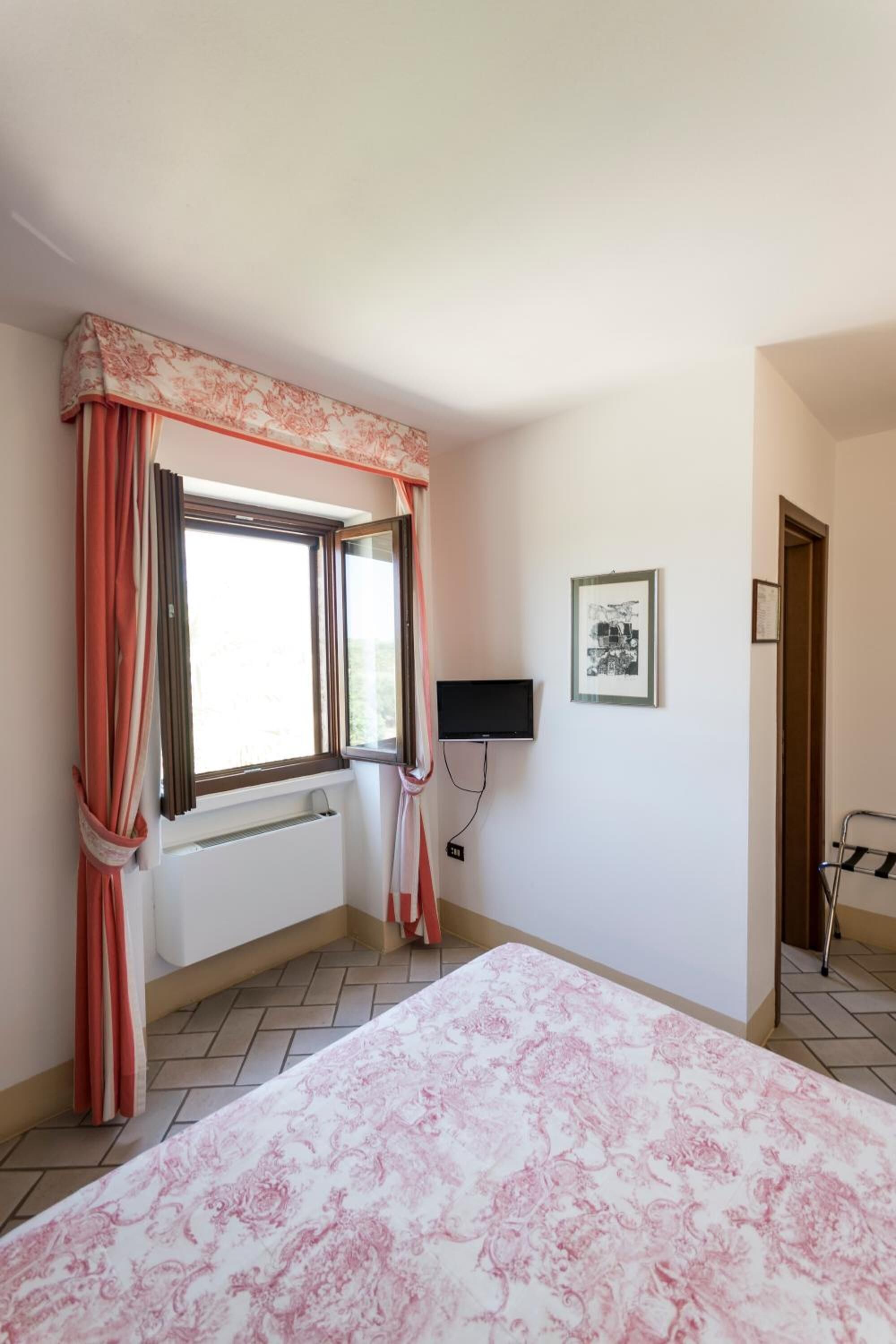 A bedroom with a bed, a window with curtains, a radiator, a television, and a framed picture on the wall.