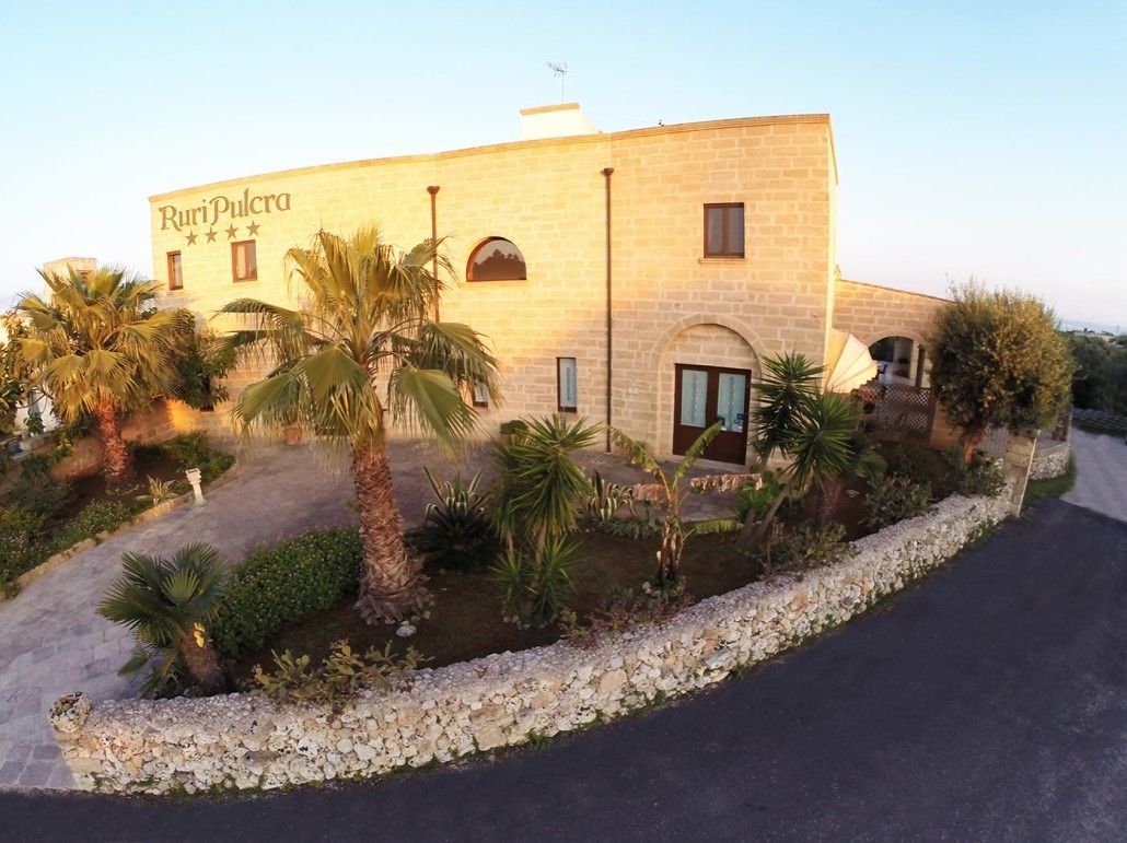 A building with the name "RuriPulcra" on the facade, surrounded by palm trees and a gravel pathway.