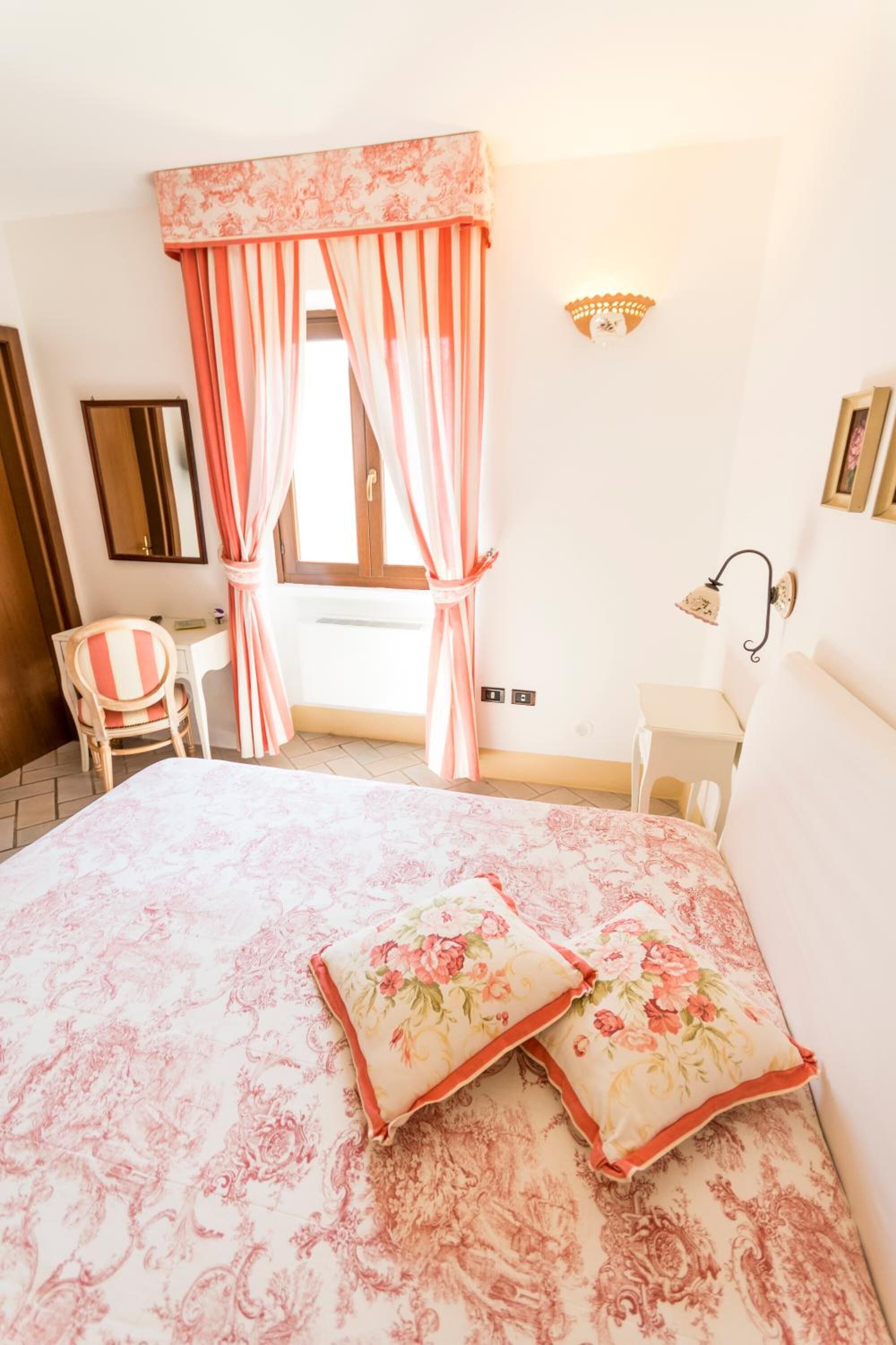 A bedroom with a bed, two pillows, a chair, a desk, a mirror, and a window with curtains.