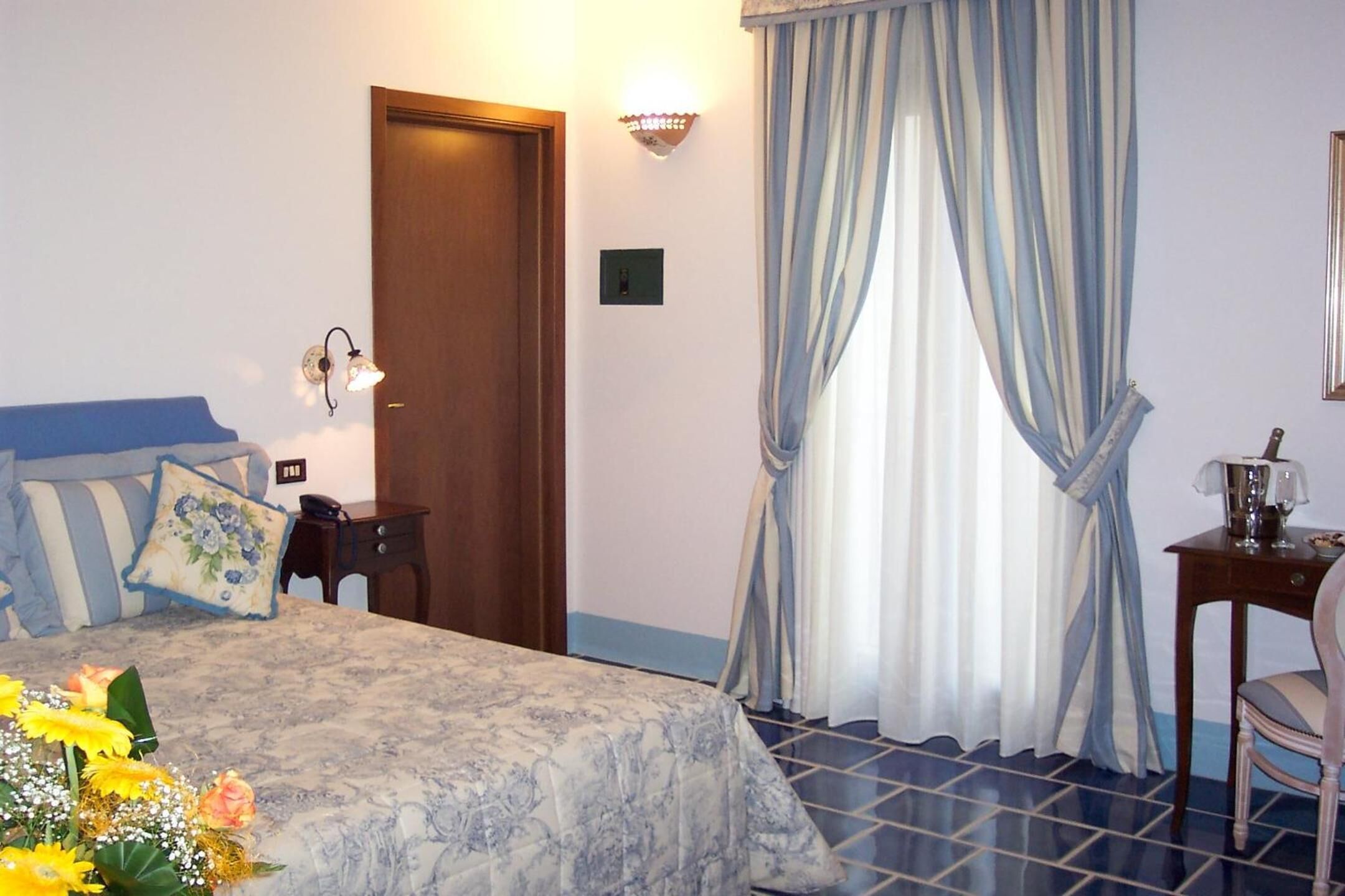 Un dormitorio con cama, escritorio, silla, mesita de noche, lámpara, cortina y puerta.
