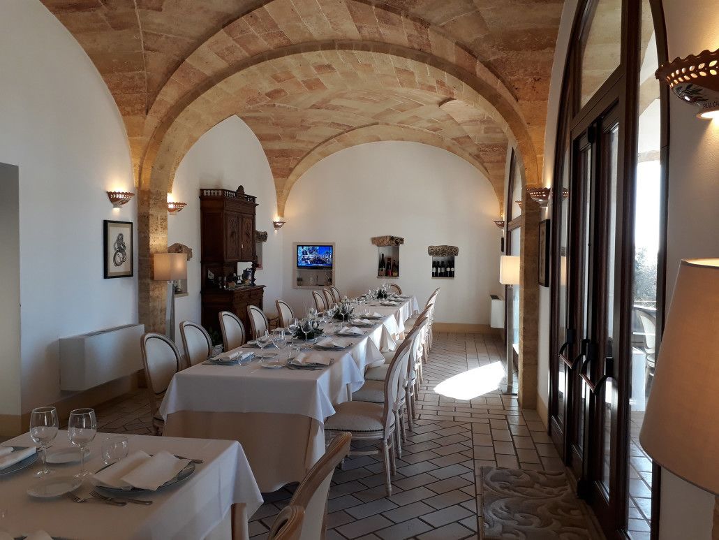 Ein langer Esstisch mit Stühlen, gedeckt für eine Mahlzeit, in einem Raum mit Rundbogenfenstern und Fliesenboden.