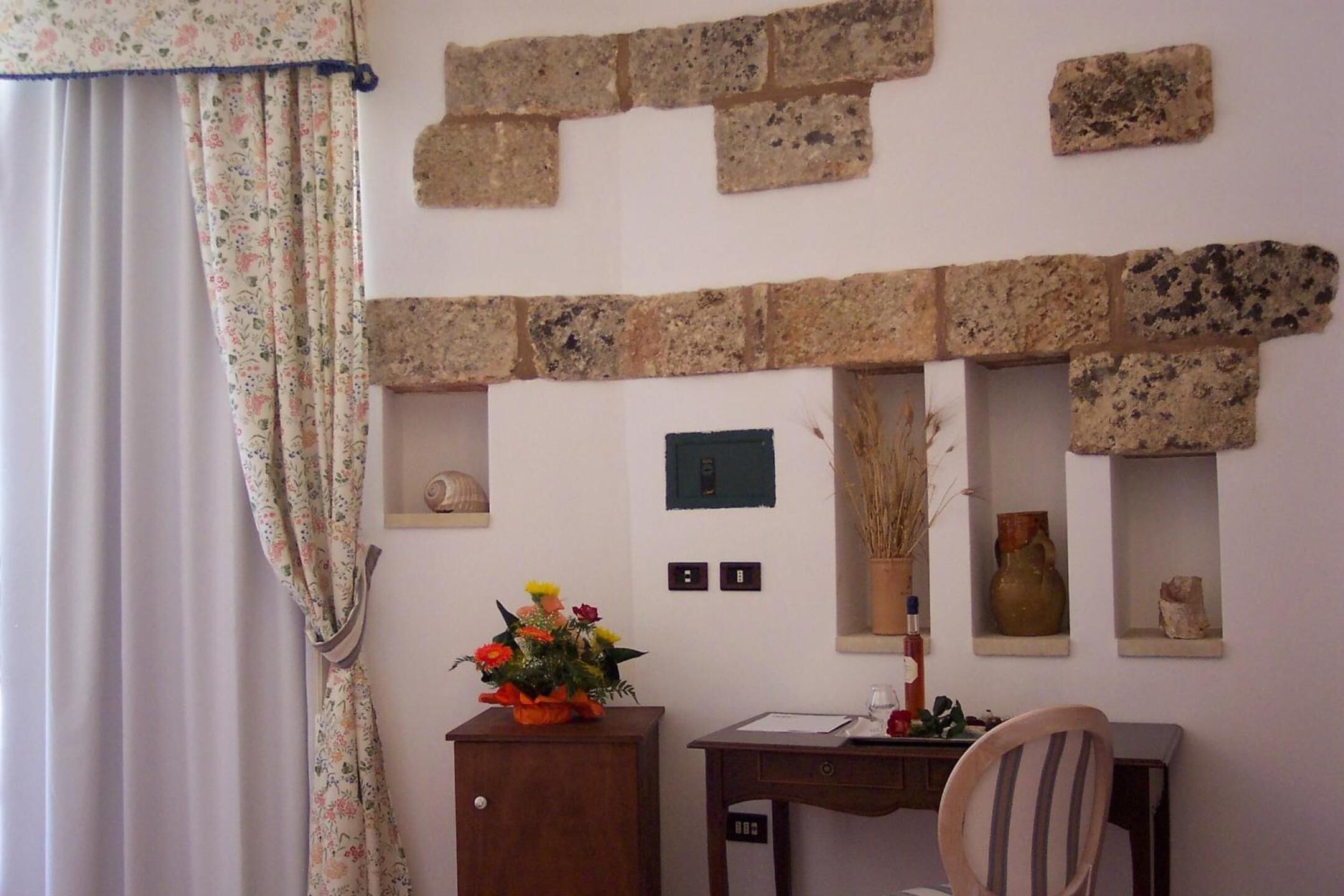 Ein Zimmer mit Backsteinwand, Schreibtisch aus Holz, Stuhl, Blumen in einer Vase und einem Vorhang.