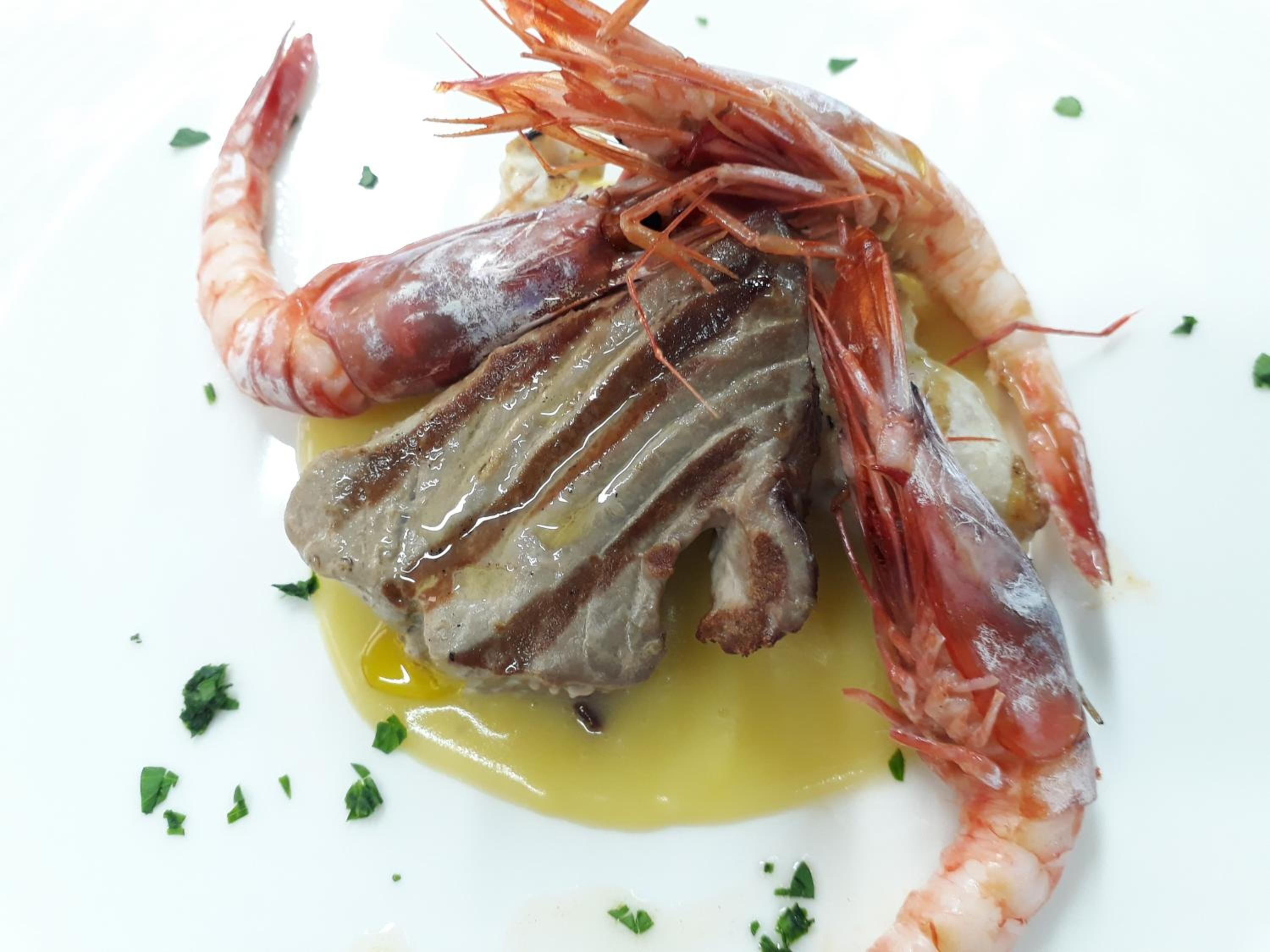 Un plato con un pescado a la parrilla, dos langostinos y una salsa amarilla, adornado con hierbas picadas.