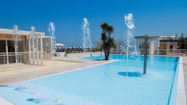 A swimming pool with multiple water jets, surrounded by a white fence and palm trees. - Hotel Aquarius (Cattolica)