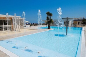 A swimming pool with multiple water jets, surrounded by a white fence and palm trees. - Hotel Aquarius (Cattolica)