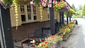 An outdoor seating area with tables and chairs, a brick wall, and hanging flower baskets. - The Flying Bull Inn (Liss)