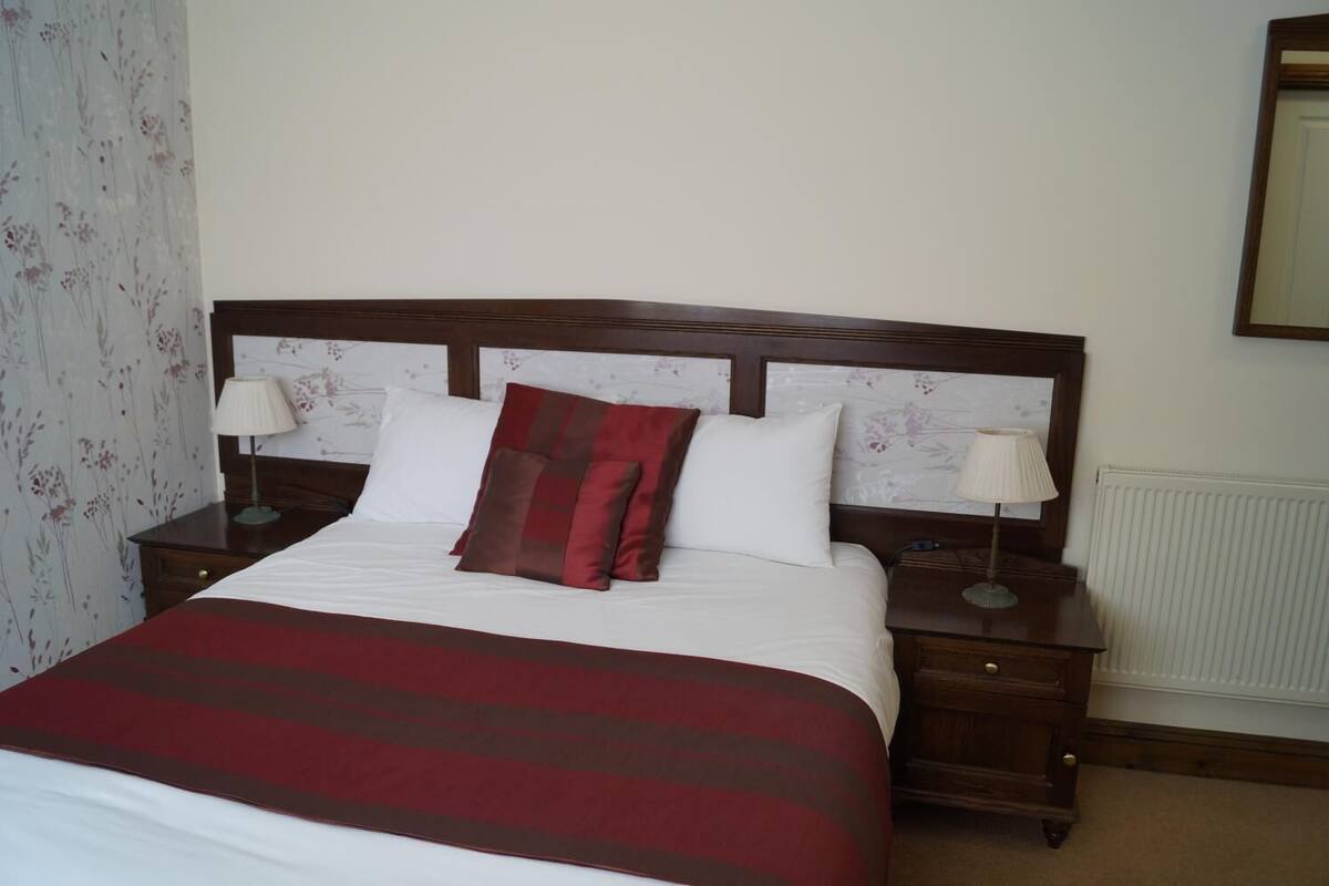A neatly made bed with a red and white striped bedspread, two white pillows, and a red and brown pillow. There are two bedside tables with lamps, a wooden headboard, and a radiator on the wall.