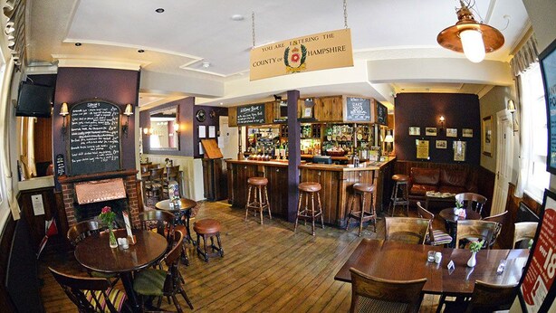 A cozy bar with wooden floors and stools, a counter with a menu, and a sign indicating the location in Hampshire. - The Flying Bull Inn (Liss)