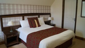 A neatly made bed with a red and white bedspread, two bedside tables with lamps, a wooden headboard, and a plain wall with a door. - The Flying Bull Inn (Liss)