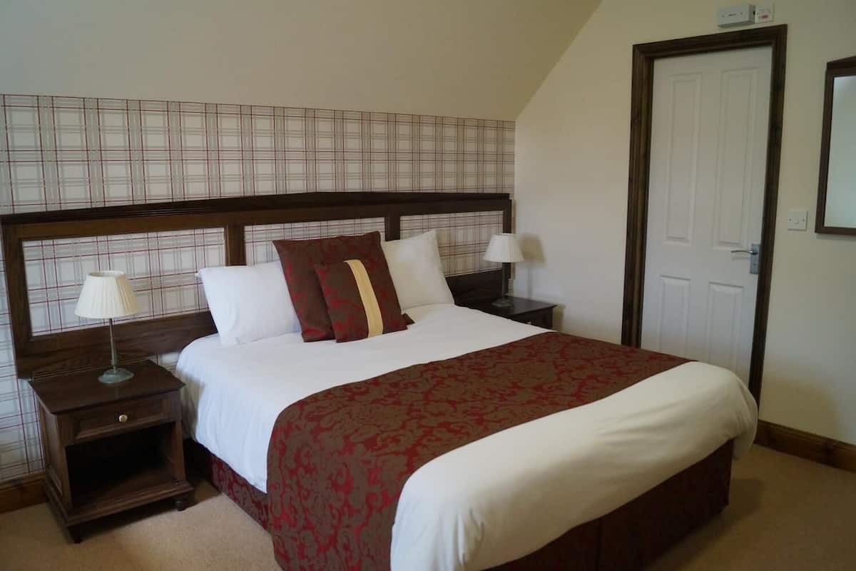A neatly made bed with a red and white bedspread, two bedside tables with lamps, a wooden headboard, and a plain wall with a door.
